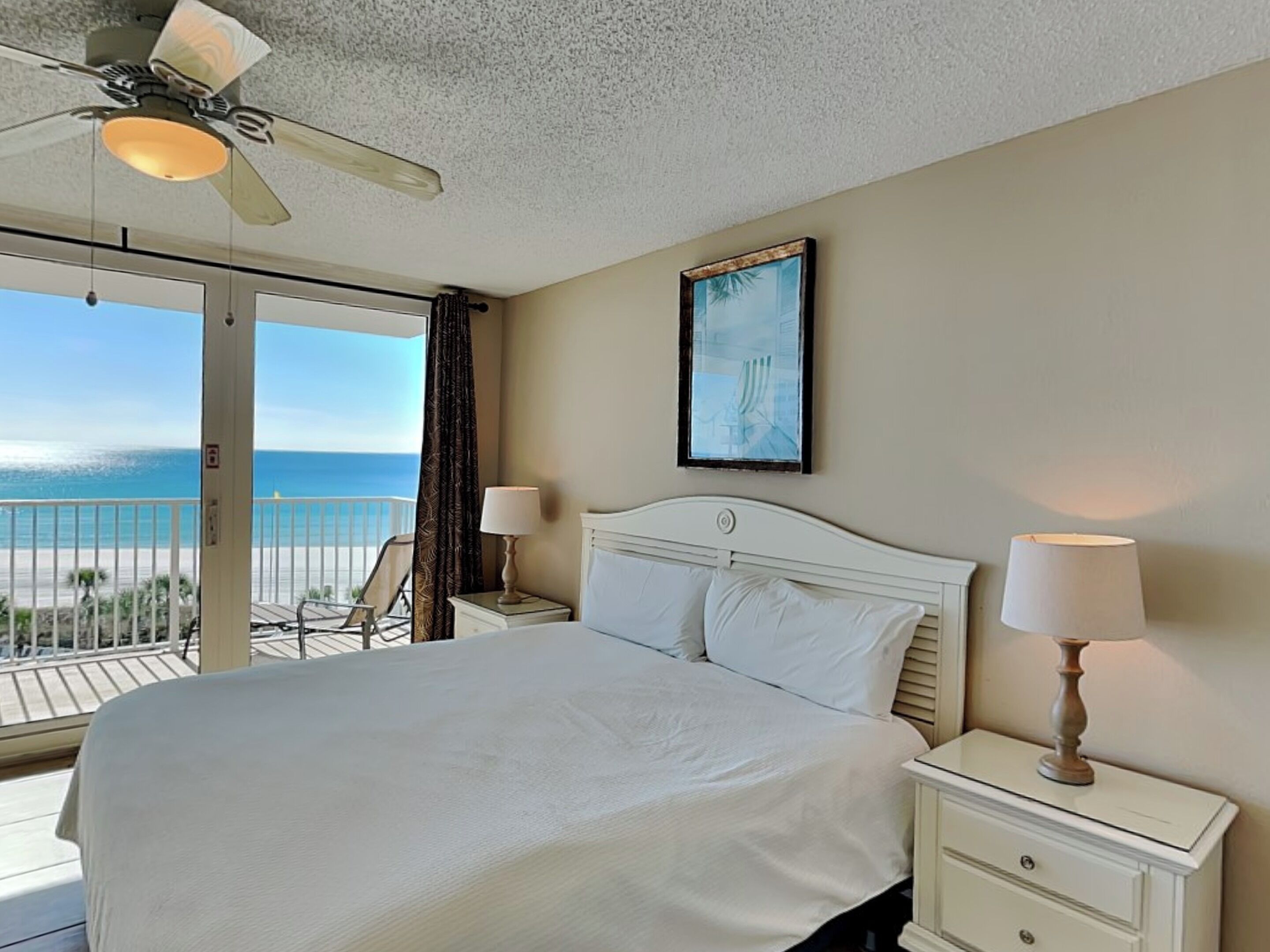 A hotel room with a large bed, two bedside tables with lamps, a ceiling fan, a painting on the wall, and a balcony with a view of the ocean.