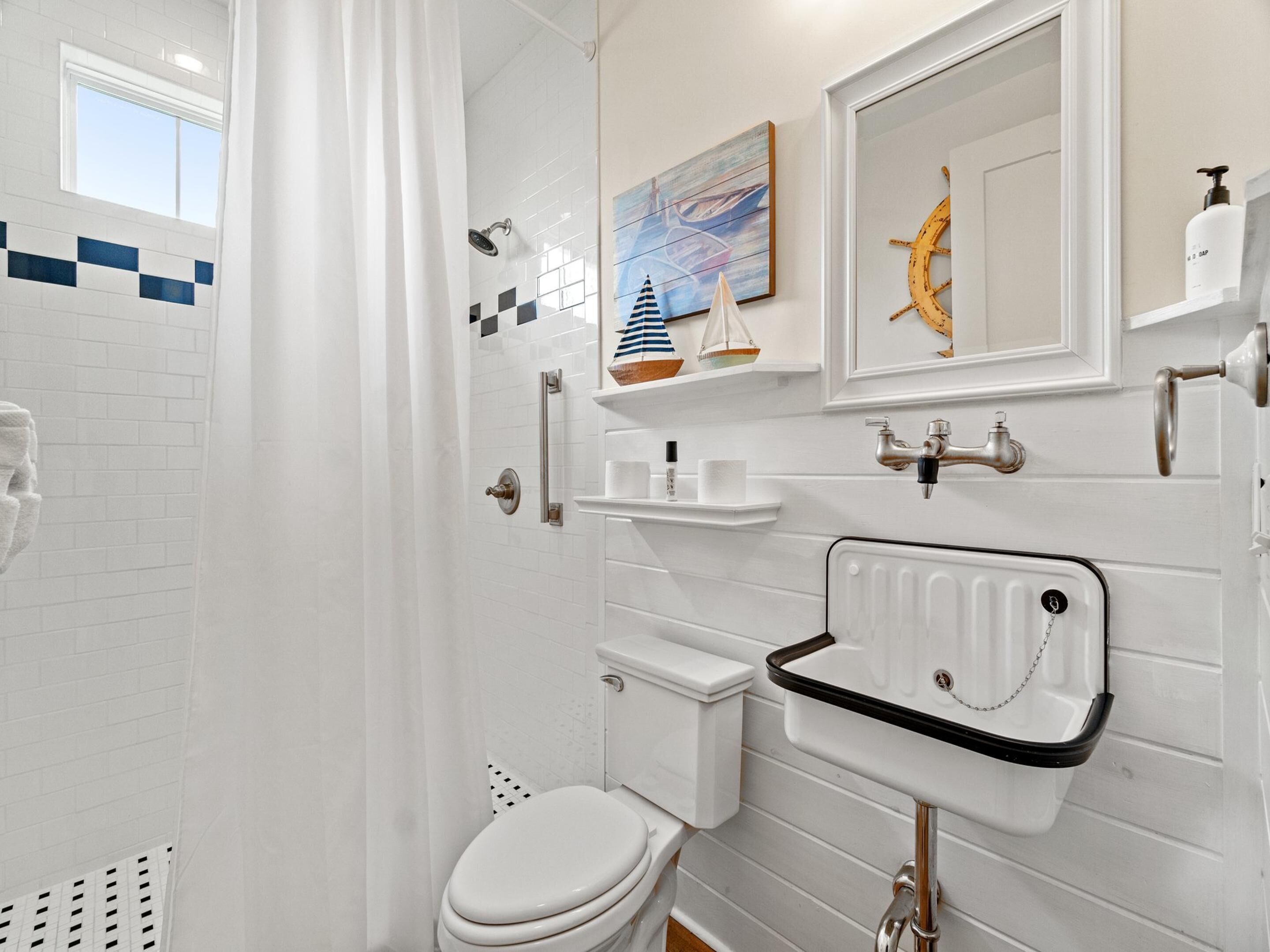 A bathroom with a white tiled wall, a white toilet, a white sink with a black countertop, a shower with a curtain, and a window with a view of the sky.