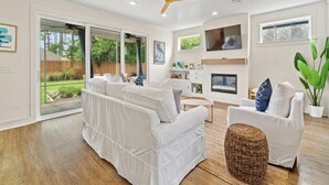 Ein geräumiges Wohnzimmer mit einem weißen Sofa, einem weißen Sessel, einem an der Wand befestigten Fernseher und einem Deckenventilator.