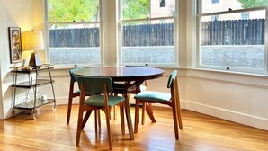 A dining area with a round table and four chairs, large windows with a view of a wooden fence and houses, and a small side table with a lamp and decorative items.