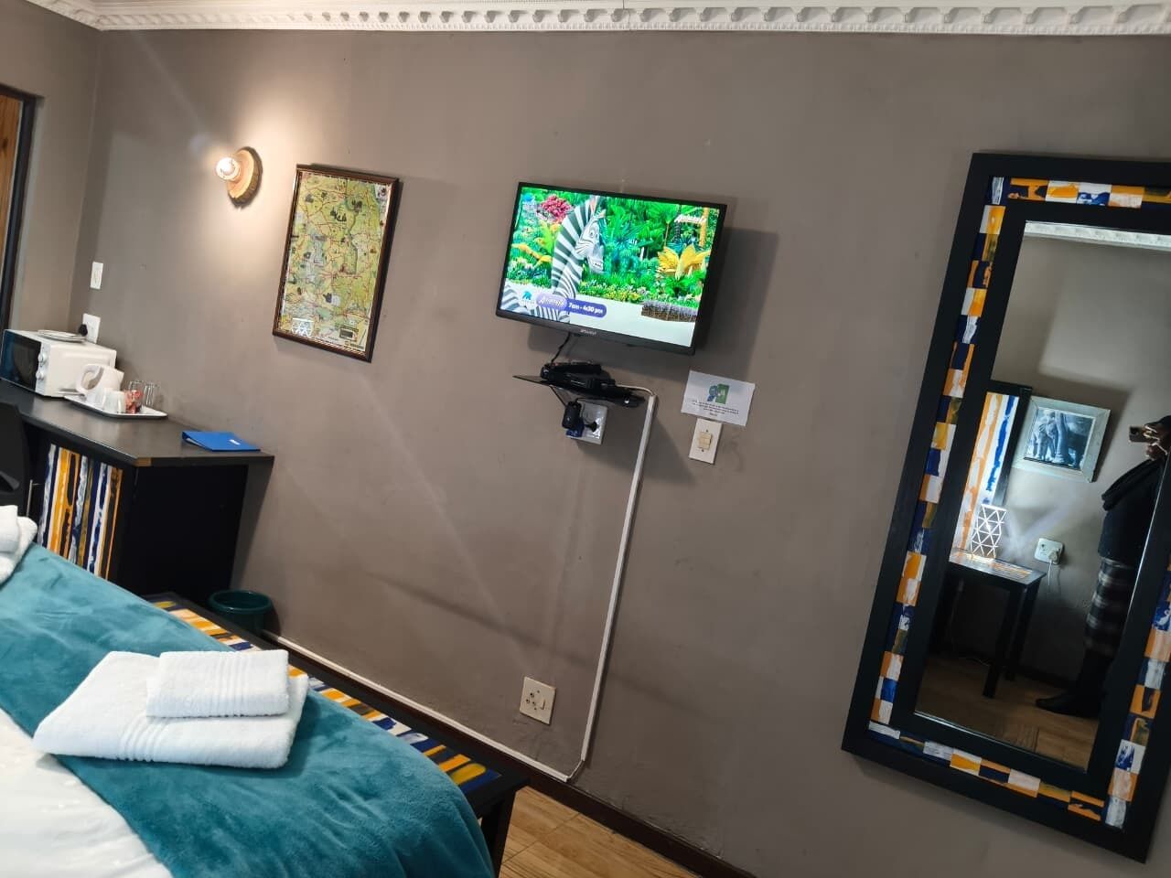 A hotel room with a bed, a television mounted on the wall, a mirror with a decorative frame, and a small table with a lamp.