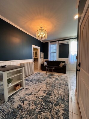 A living room with a dark blue wall, a chandelier, a black sofa, a white radiator, and a patterned rug. - 210 Main St (Ellsworth)