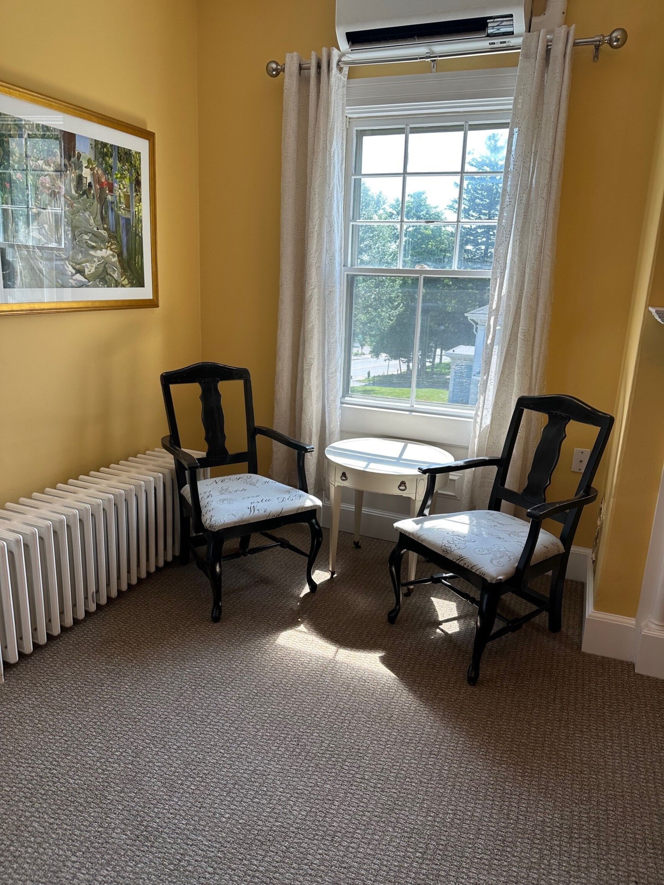 Une pièce avec deux chaises noires dotées de coussins à motifs, une petite table blanche, un radiateur et une fenêtre avec des rideaux blancs.