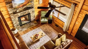 A cozy cabin interior with a stone fireplace, a ceiling fan, a comfortable sofa, and a television mounted above the fireplace.