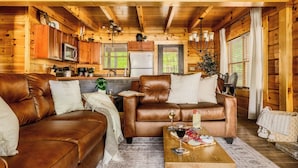 A cozy cabin interior with a living room featuring two brown leather sofas, a coffee table with wine glasses, and a kitchen area with wooden cabinets and a refrigerator.