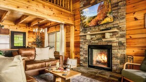 A cozy cabin interior with a stone fireplace, leather furniture, and a large window overlooking a forest.