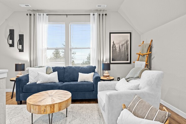 Ein modernes Wohnzimmer mit einem blauen Sofa, einem weißen Sessel, einem runden Holztisch und einem gerahmten Bild an der Wand.