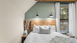 Un dormitorio con techo inclinado, cabecera de madera, dos lámparas de noche, una ventana con persianas y una mesita de noche con una lámpara y una planta.
