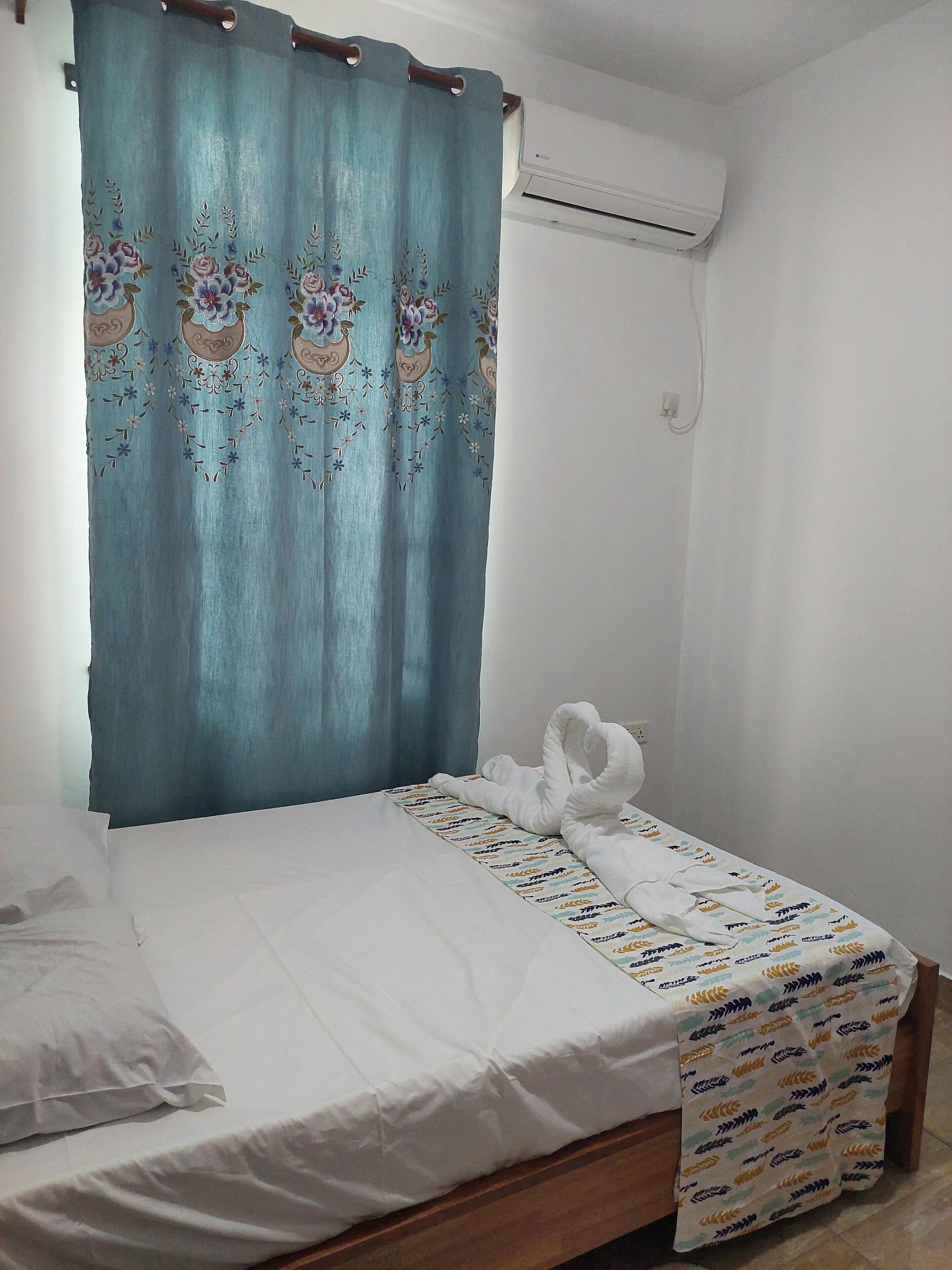 Een enkel bed met een bedovertrek met dessin, een blauw gordijn met bloemmotieven, en een airconditioning op de muur.
