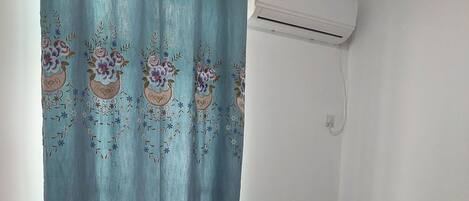 Een enkel bed met een bedovertrek met dessin, een blauw gordijn met bloemmotieven, en een airconditioning op de muur.