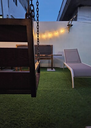 Un patio con una hamaca, dos sillones y una guirnalda de luces frente a una pared blanca.