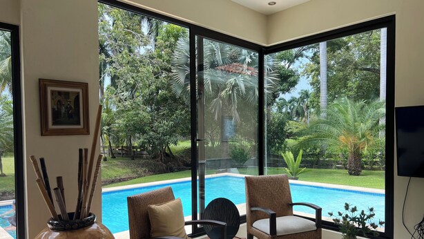 Quarto com uma grande janela com vista para uma piscina e vegetação exuberante. Há duas cadeiras de vime com almofadas, uma mesa de madeira e um vaso decorativo.