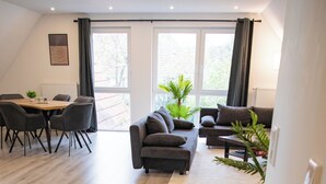 Un salón amplio con un conjunto de sofás, una mesa de comedor y sillas. Se observan ventanas grandes con cortinas, y un radiador en la pared.