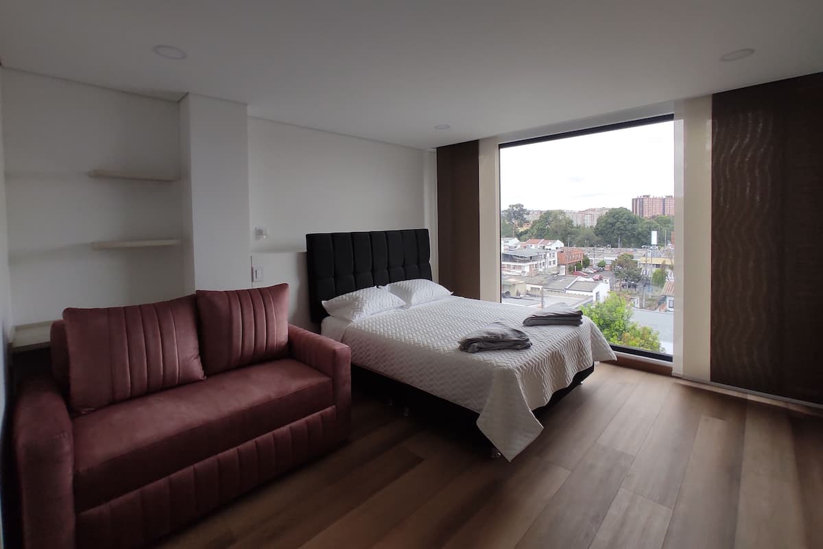 Quarto moderno com cama, sofá vermelho e uma grande janela com vista para a cidade.