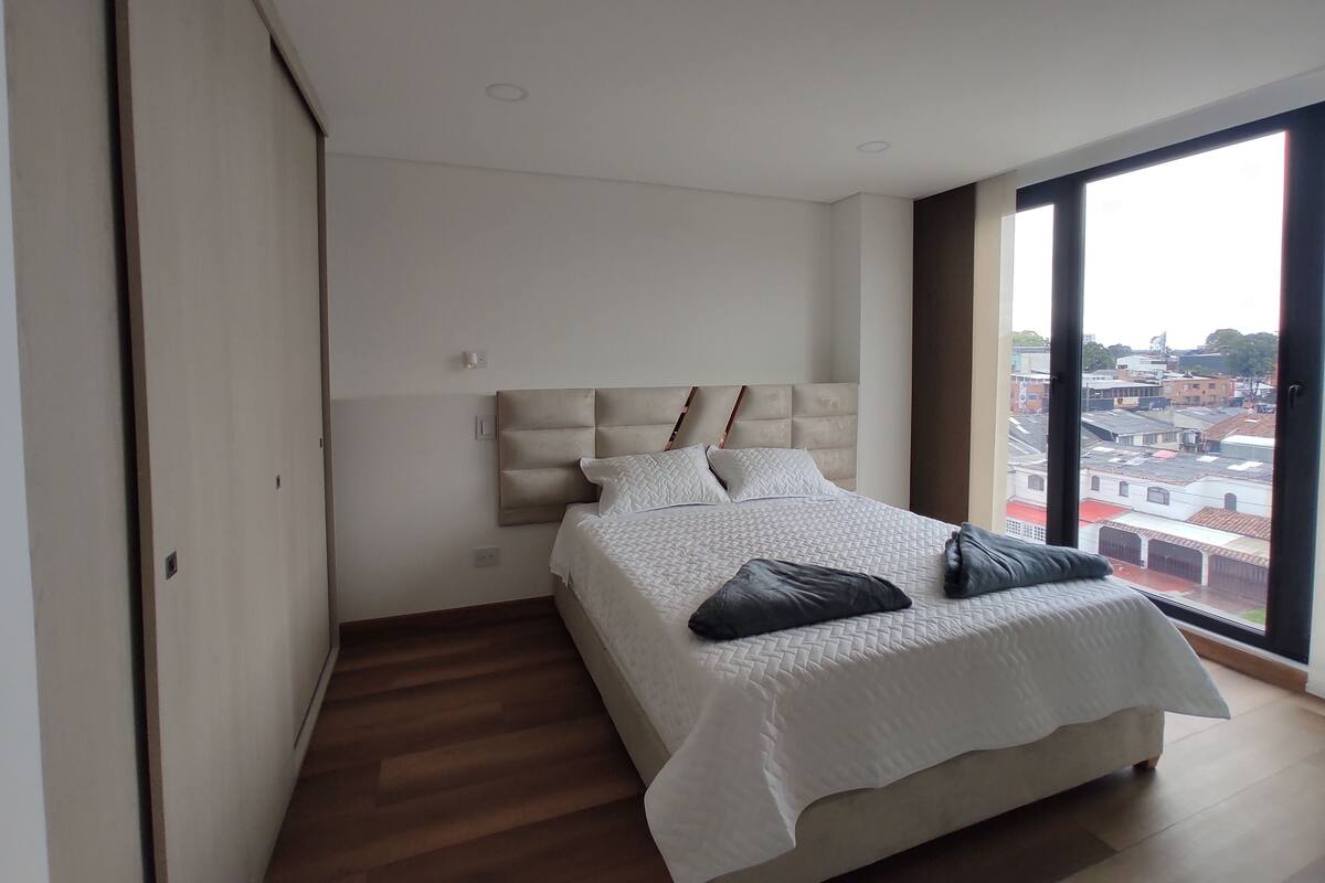 Quarto moderno com uma cama grande, cabeceira com compartimentos de armazenamento, vista da cidade através de uma janela ampla e piso de madeira.