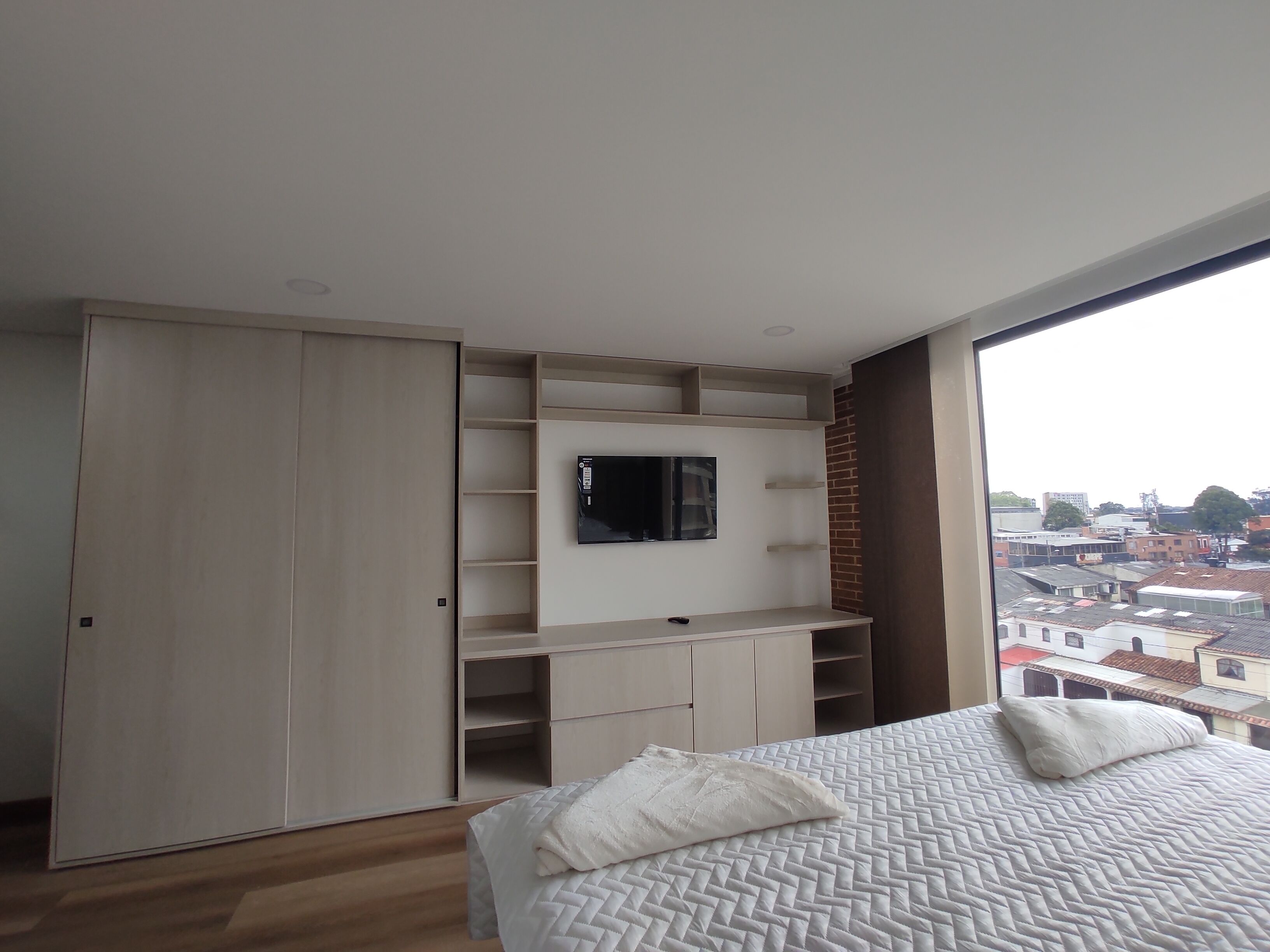 Een slaapkamer met een bed, een aan de muur bevestigde televisie en uitzicht op de stad door een groot raam.