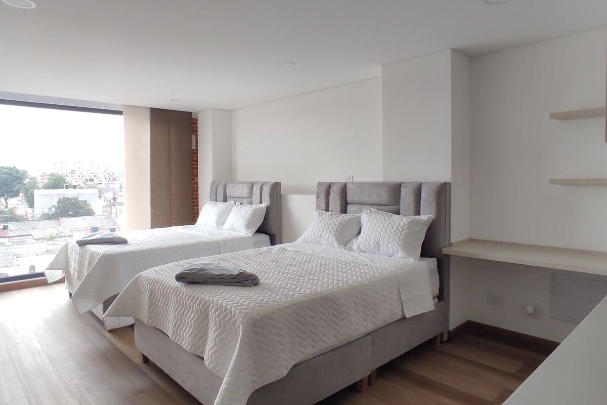 Quarto de hotel moderno com duas camas, uma janela grande e vista para a cidade.