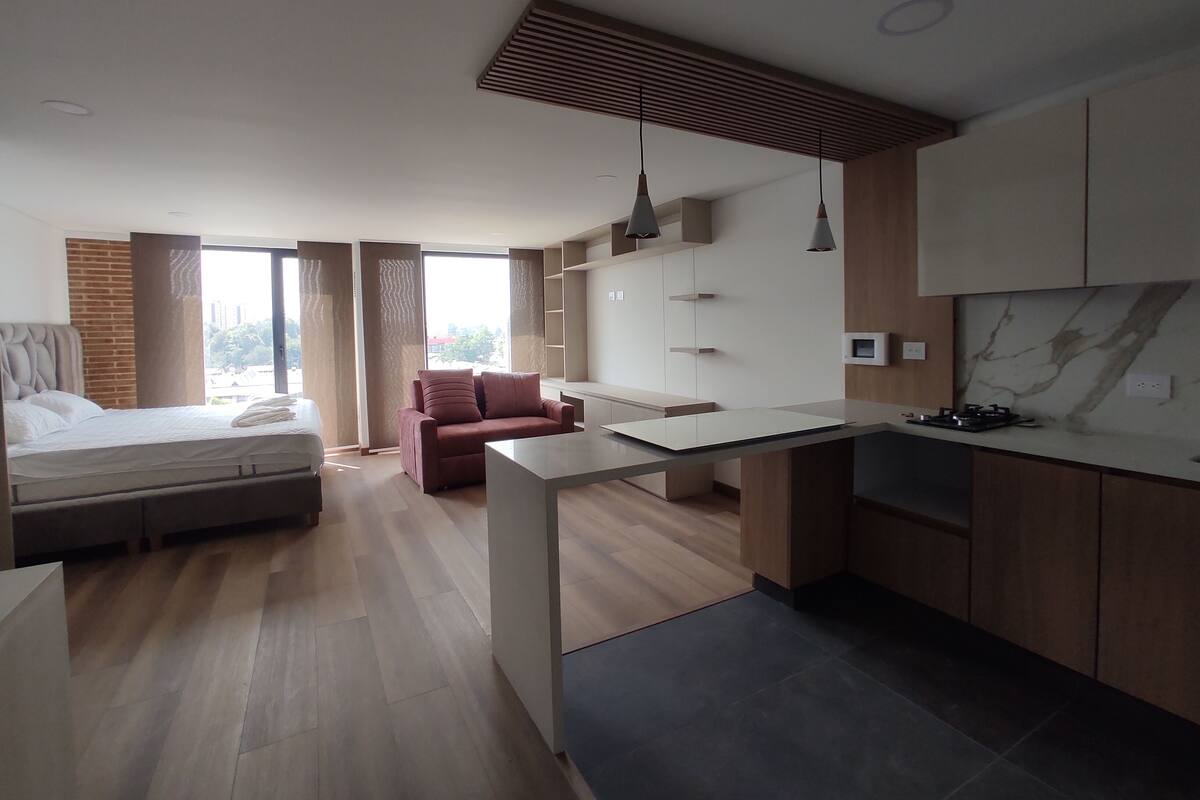 Quarto de hotel moderno com cama, sofá, pequena cozinha e vista para a cidade.