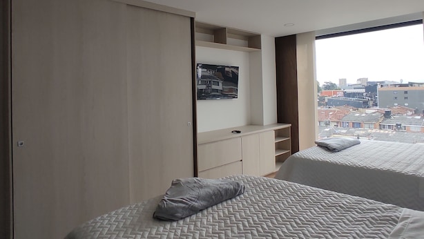 A modern bedroom with a large window, a bed with a grey pillow, and a wardrobe. - Modelia Living & Parking (Bogotá)