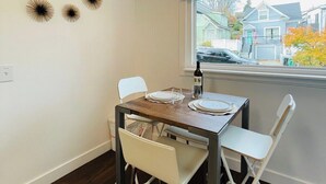 Un coin repas avec une table dressée pour deux personnes, une fenêtre donnant sur des maisons et un luminaire au plafond.