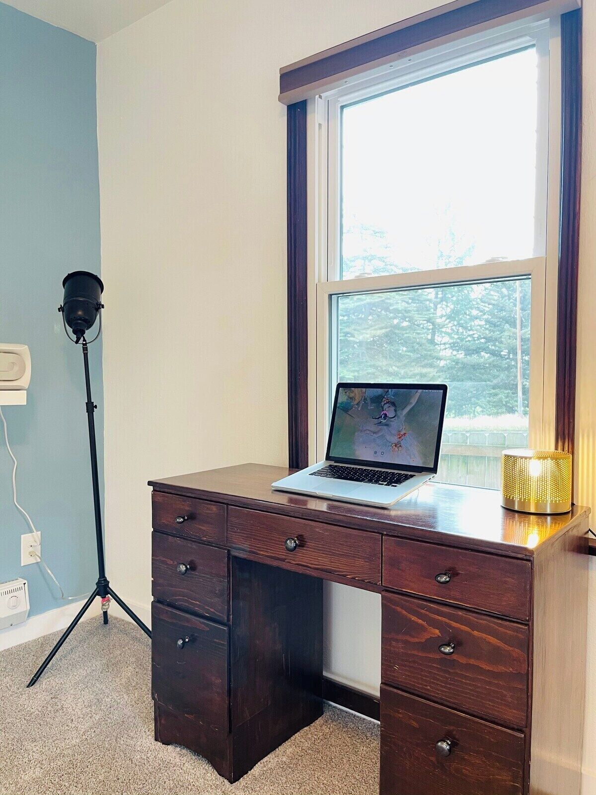 Un bureau en bois avec un ordinateur portable, une lampe sur pied et une fenêtre donnant sur des arbres.