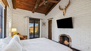 Une chambre avec un mur en briques blanches, une cheminée, un lit, une télévision et une porte donnant sur un balcon.
