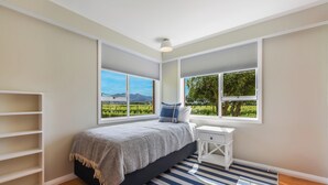 A bedroom with a bed, a nightstand, a built-in shelf, and large windows with a view of a vineyard. - Springfields Homestead - Rapaura Holiday Home (Blenheim)
