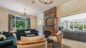 A living room with a brick fireplace, dark green sofas, and a large window with a view of greenery. - Springfields Homestead - Rapaura Holiday Home (Blenheim)