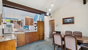 A kitchen with wooden cabinets, a sink, and a countertop. There is a dining area with a table and chairs. A framed picture is on the wall. - Sunlit Solace - Wanaka Holiday Home (Wanaka)