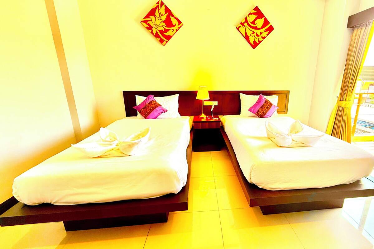 A hotel room with two beds, a nightstand, a lamp, and a view of a building through the window.