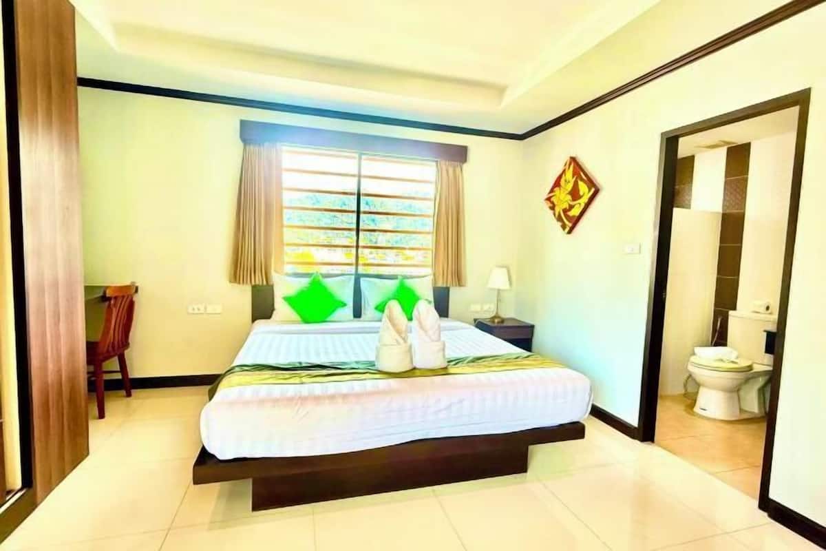 A hotel room with a bed, a desk, a chair, a window with curtains, a nightstand, a lamp, and a bathroom visible through an open door.
