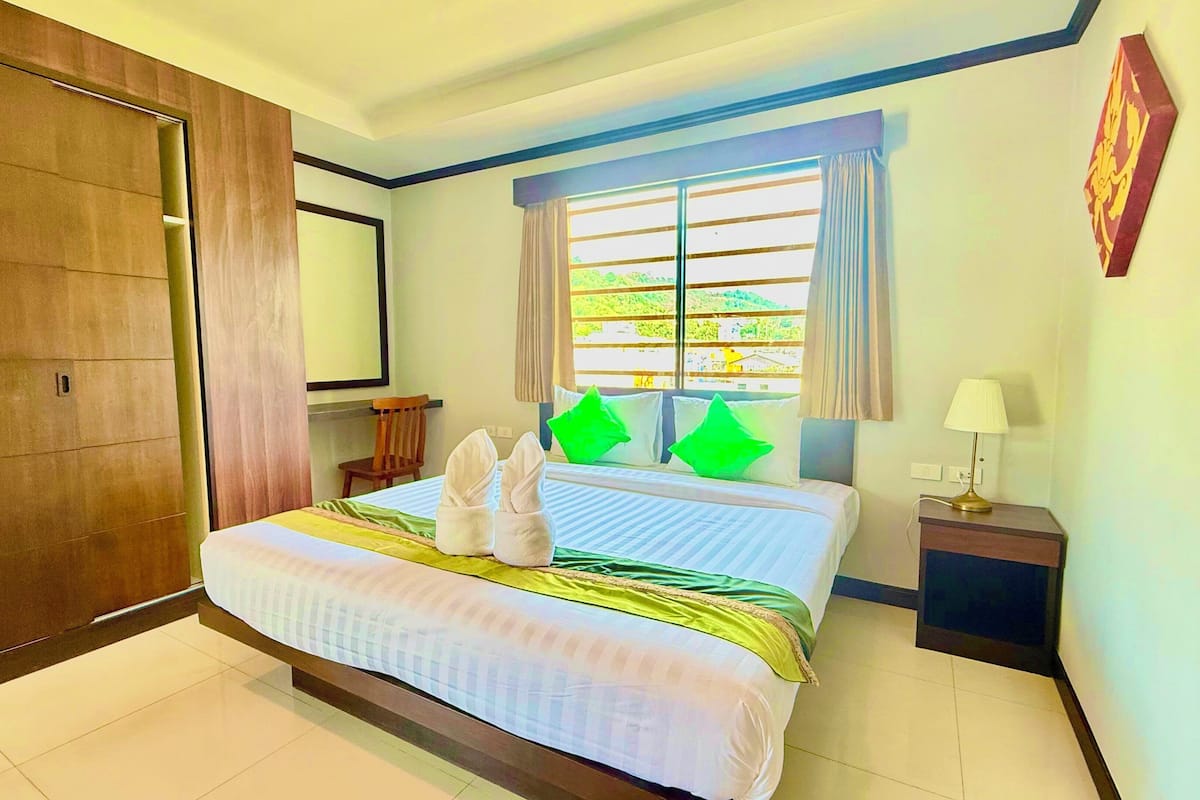 A hotel room with a large bed, a wooden wardrobe, a window with curtains, a bedside table with a lamp, and a wall-mounted artwork.