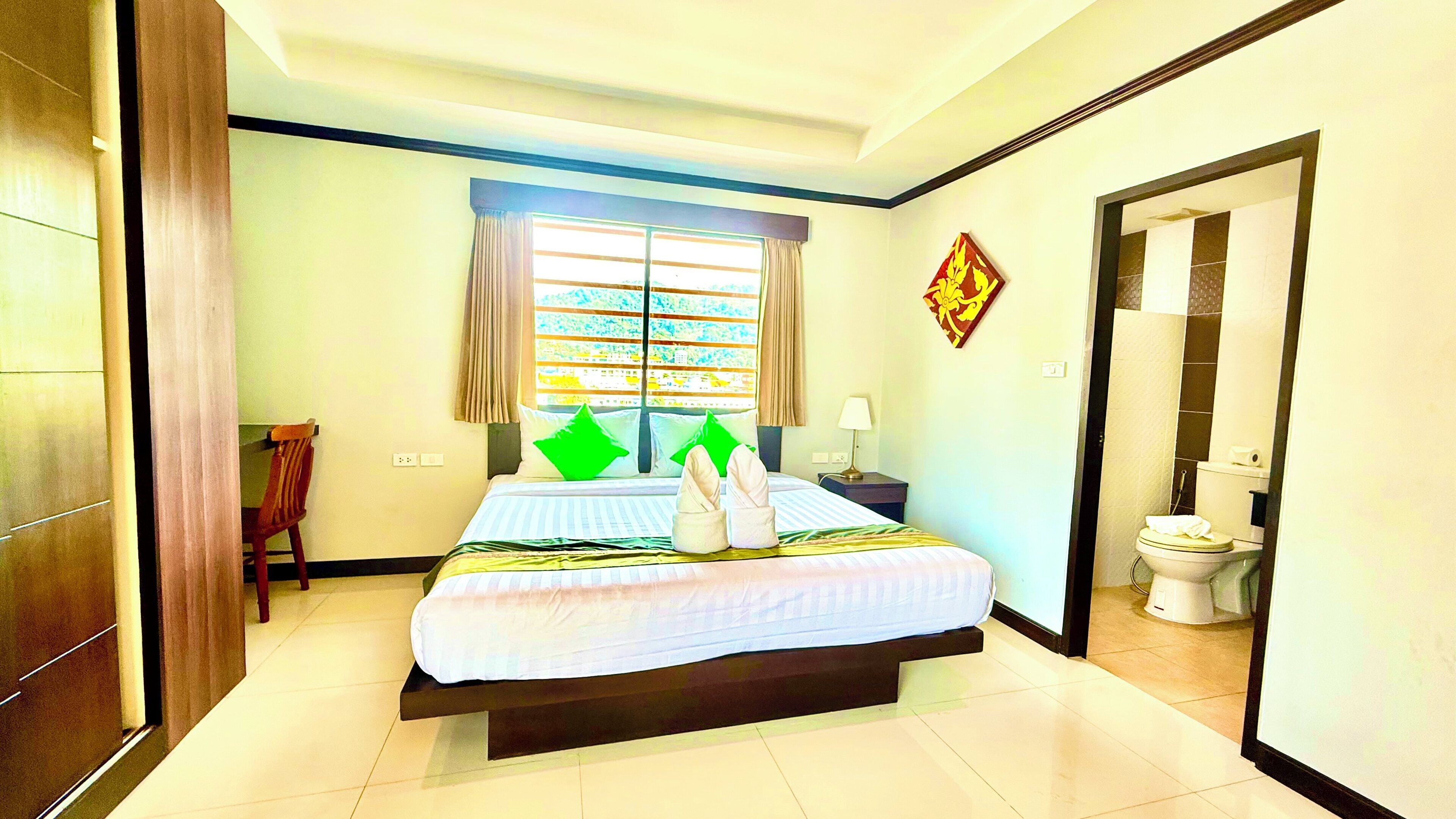 A hotel room with a bed, a window with curtains, a nightstand, a chair, and a bathroom visible through an open door.