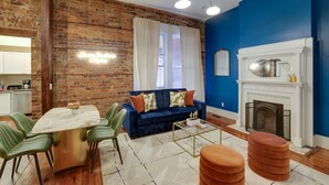 Een eetgedeelte met een marmeren tafel en groene stoelen, een blauwe bank met oranje kussens en een witte schoorsteen.