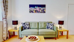 A living room with a green sofa, two side tables with lamps, a painting on the wall, and a bowl of fruit on the coffee table.