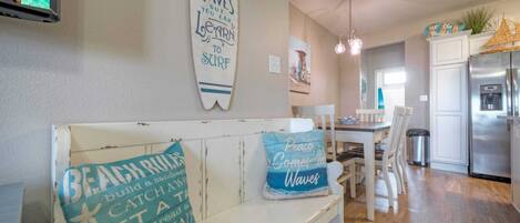 Un salon chaleureux avec un banc blanc, une décoration murale représentant une planche de surf et une cuisine en arrière-plan.