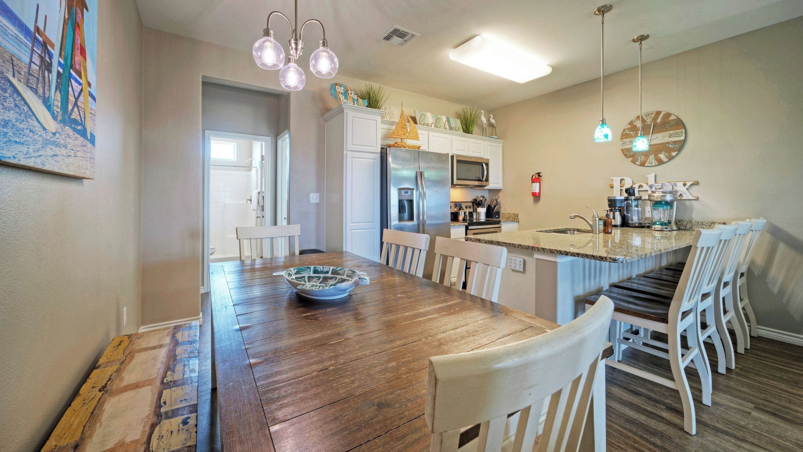 A kitchen with a wooden dining table, white chairs, a refrigerator, and a coffee maker.
