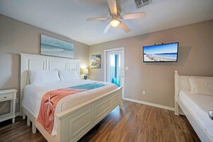 A bedroom with a large bed, a ceiling fan, a television, and a door leading to a balcony.