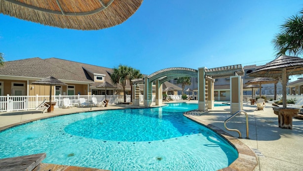 A swimming pool with a white deck, a covered seating area, and a building with a tiled roof in the background. - The Beautiful Place by Avantstay Mins to Beach + Family-friendly Pool (Corpus Christi)
