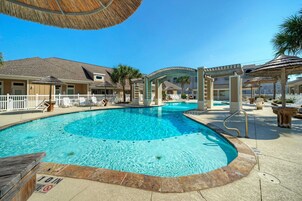 A swimming pool with a white deck, a covered seating area, and a building with a tiled roof in the background.