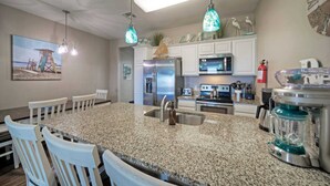 A kitchen with granite countertops, white cabinetry, a large island with seating, pendant lights, and a beach-themed painting. - The Beautiful Place by Avantstay Mins to Beach + Family-friendly Pool (Corpus Christi)