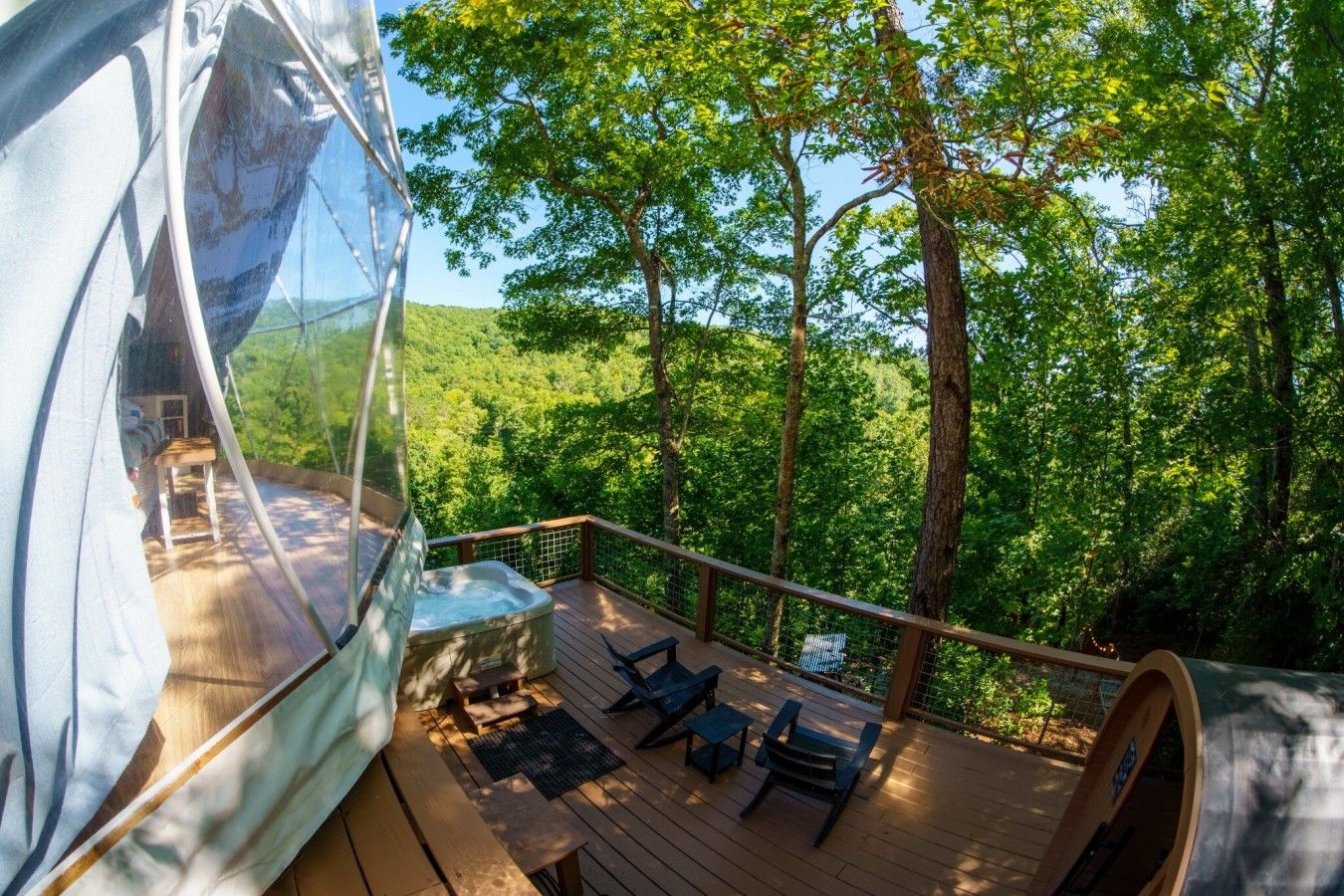 Une structure en forme de dôme transparent, dotée d’une terrasse en bois, de sièges extérieurs et offrant une vue sur un paysage boisé.