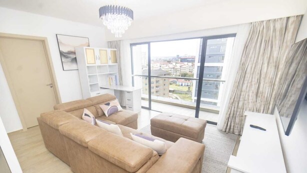 Un salon moderne avec un canapé d’angle beige, un meuble blanc et une grande fenêtre donnant sur la ville.