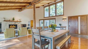 Un vaste salon doté d’une cheminée en pierre, de poutres apparentes en bois et de grandes fenêtres offrant une vue sur les arbres.