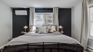 A bedroom with a bed, bedside lamps, a window with curtains, and an air conditioning unit. - Dunes & Waves Retreat -  Perfect Lake Getaway (Mears)