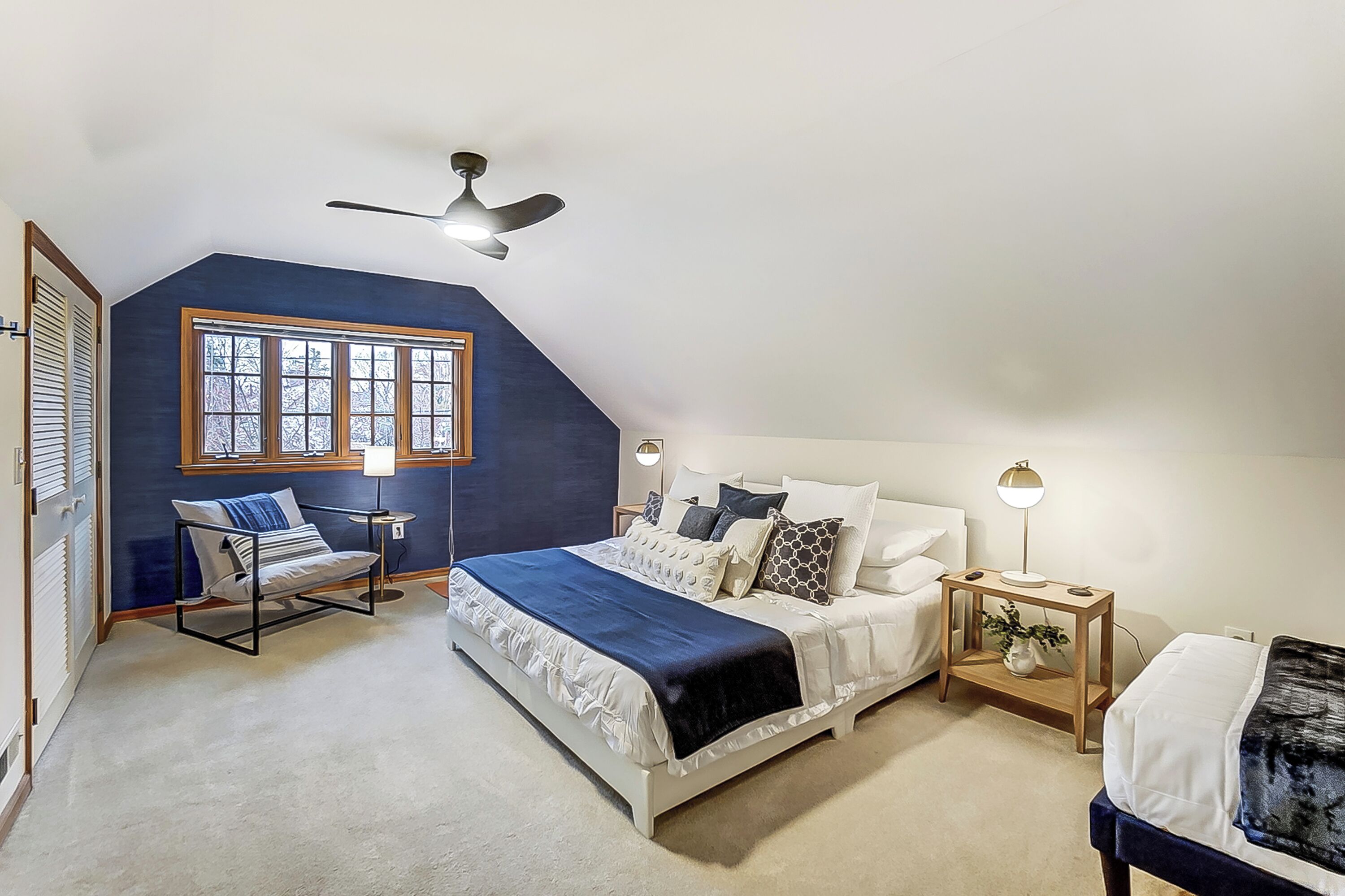 A bedroom with a sloped ceiling, a large bed with multiple pillows, a side table with a lamp, a chair, and a window with blinds.