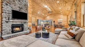 A log cabin interior with a stone fireplace, wooden ceiling, and a cozy living area with sofas and a dining table.