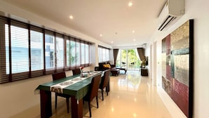 A dining area with a long table, chairs, and a sofa. There are large windows with blinds, a ceiling fan, and a view of greenery outside.
