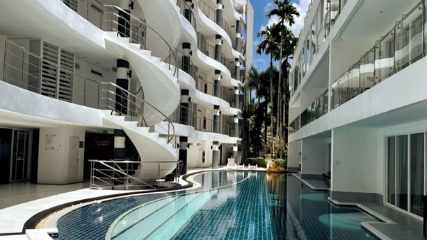 A modern hotel with a central swimming pool, multiple balconies, and a tropical garden area.