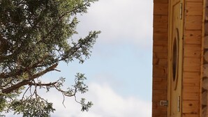 Een houten hutje met een deur en een raam, een boom op de voorgrond en een bergachtig landschap op de achtergrond.